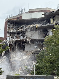 Partially demolished concrete building with debris and dust clouds beside green trees