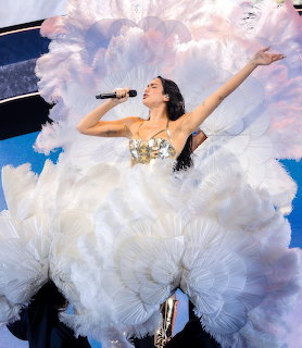 Dua Lipa singing on stage in a voluminous white ruffled dress with one arm raised
