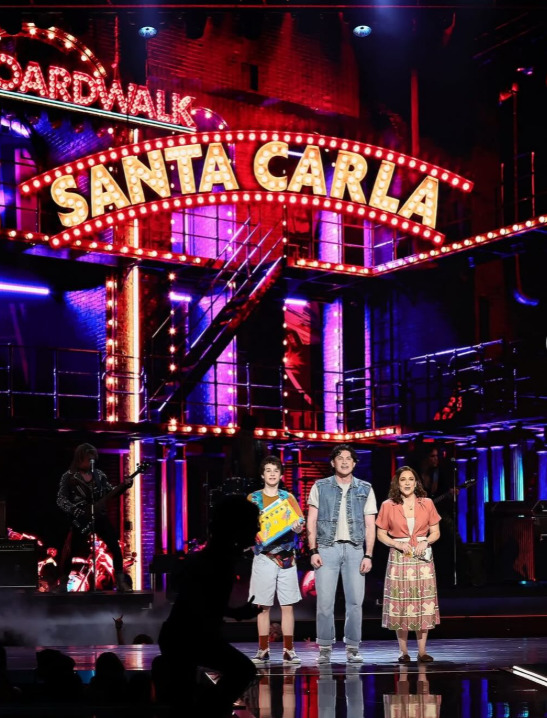 Four people standing on stage under bright Sardi's marquee with colorful theatrical lighting