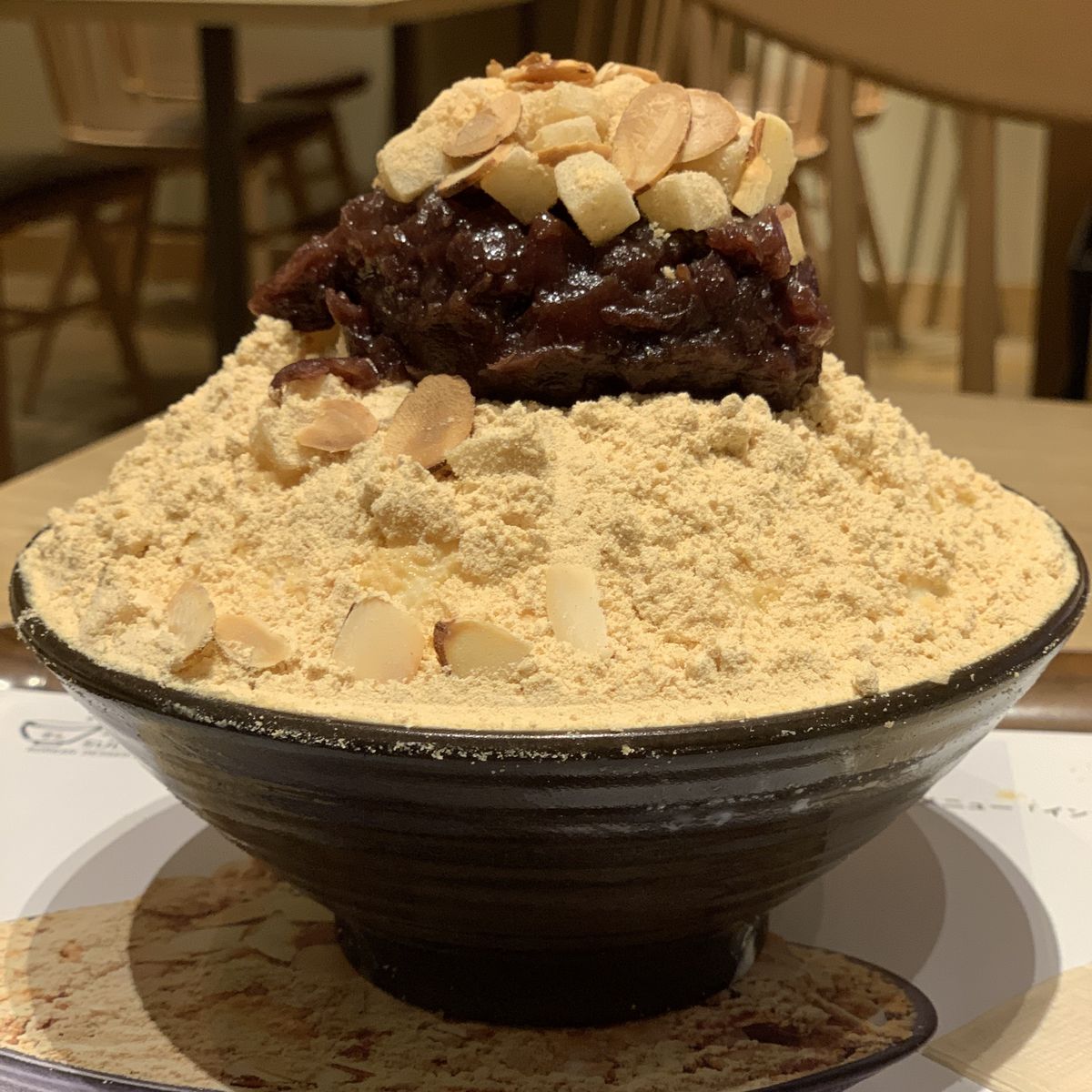 Bowl of shaved ice mounded with tan kinako powder, red bean paste topping, and chopped nuts in indoor cafe setting