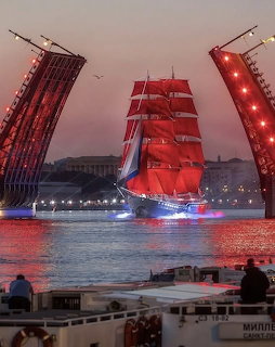 Tall ship with red sails passing through an open drawbridge at night with city lights
