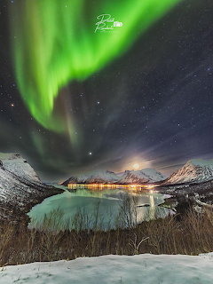 Green northern lights over snowy mountains and fjord with lights from a small village in Senja, Norway