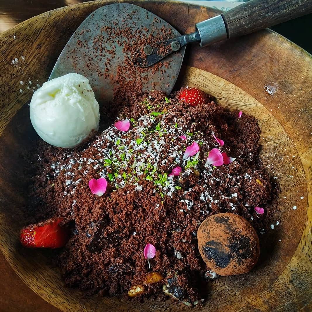 Chocolate dessert styled as dirt pile with flowers, ice cream, strawberry, and small shovel on wooden plate