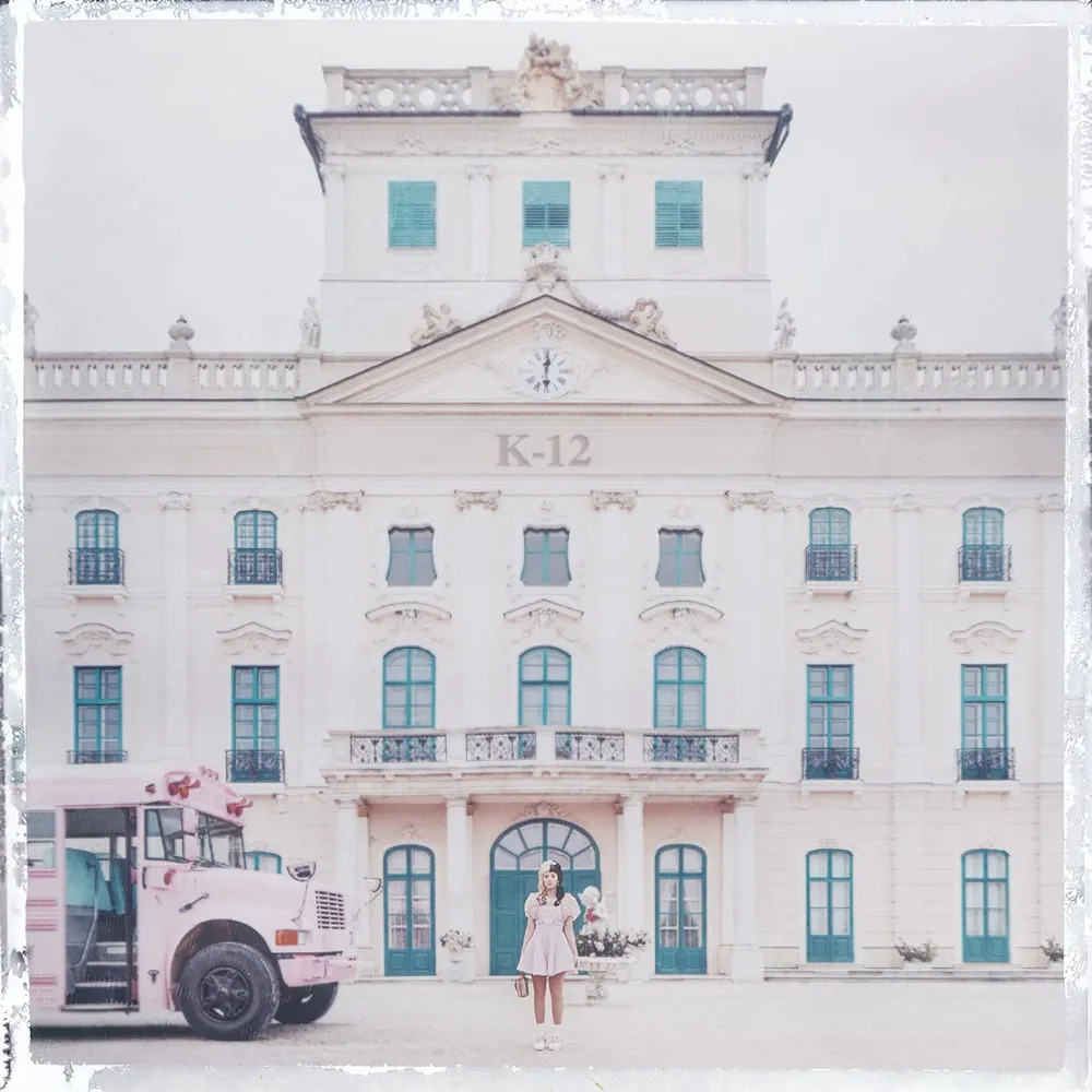 Pastel neoclassical school building with K-12 text, figure at entrance, pink bus on left