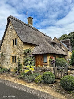 Stone cottage with thick thatched roof, colorful front garden, and rustic wooden fence