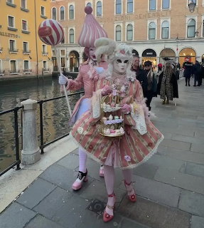 Person in elaborate pink Venice carnival costume with mask holding tiered sweets display by canal