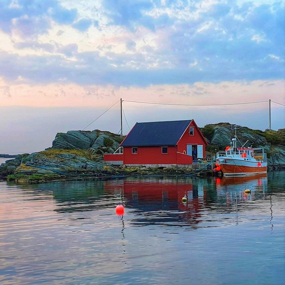 Red fishing shack and boat reflected in calm coastal water beside rocky shore at dusk