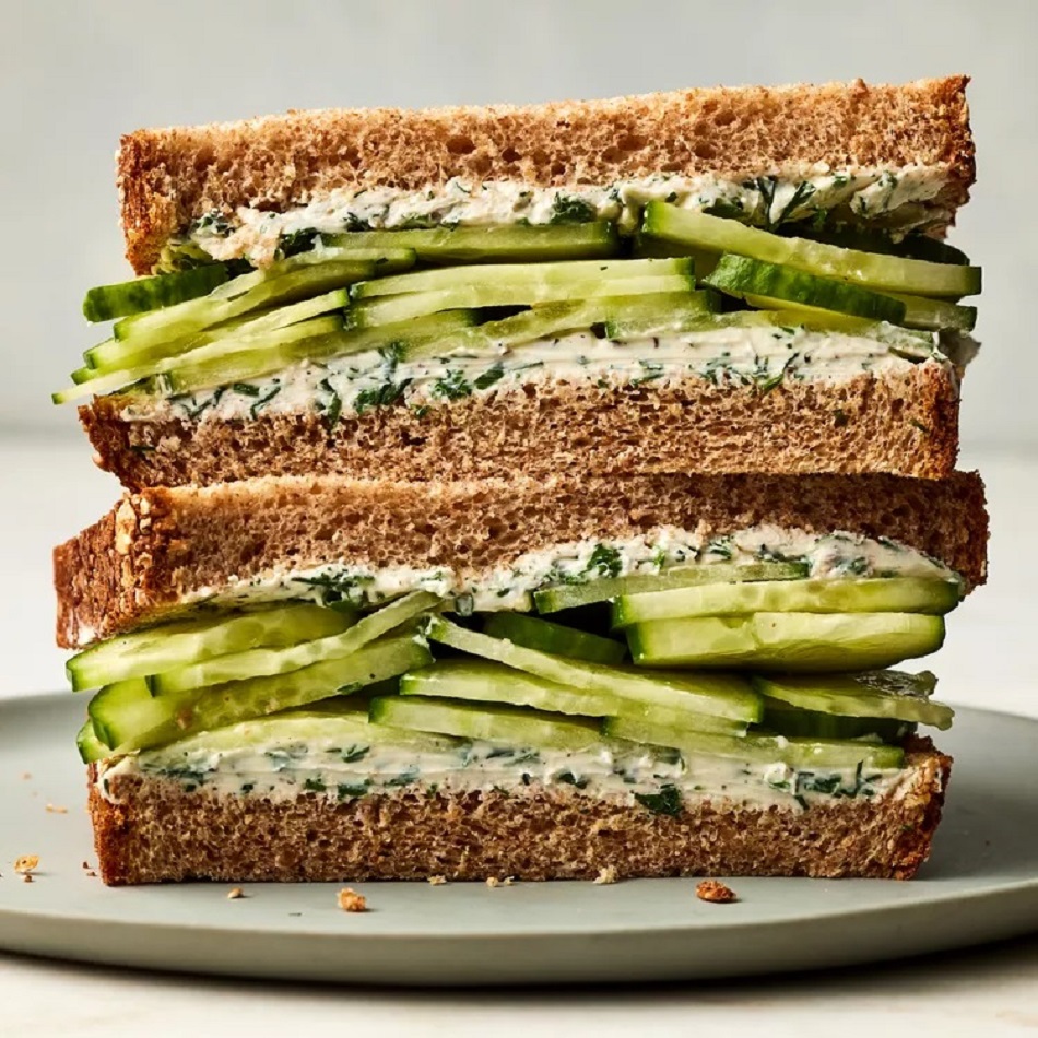 Two stacked cucumber sandwich halves on wheat bread with cream cheese and fresh cucumber slices on a white plate