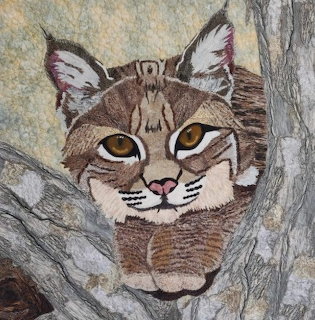 Fabric quilt artwork of a bobcat with amber eyes resting among gray tree branches