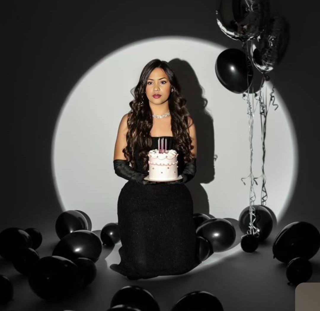 Woman in a black dress holding a cake under a spotlight with black balloons around her
