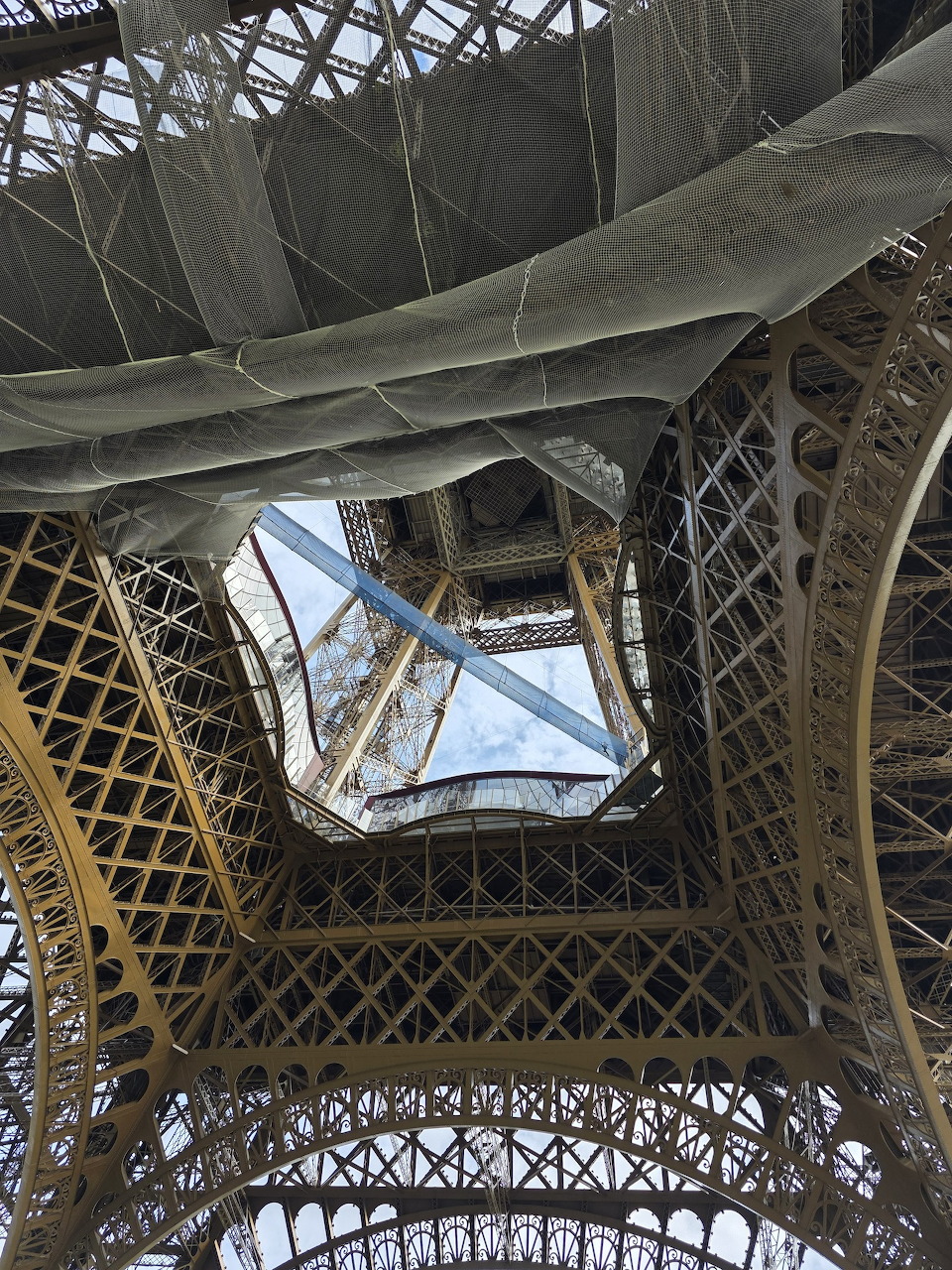 Looking up through the Eiffel Tower's iron lattice structure toward a glass ceiling and blue sky, Paris