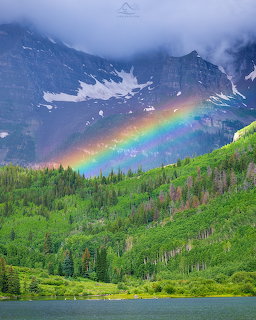 Rainbow arcing over Maroon Bells mountains with green aspen forest and alpine lake in foreground