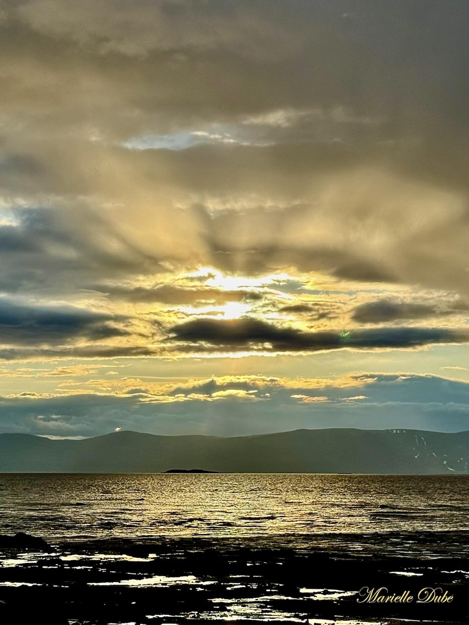 Golden sunset over calm water with distant mountains, dramatic clouds, and a dark rocky shoreline