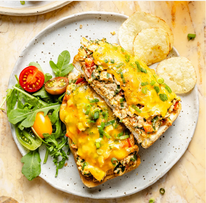 Open-faced chicken melts with melted cheese, salad, cherry tomatoes, and potato chips on a plate