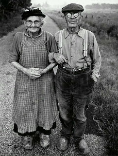Black and white portrait of elderly couple in rural setting, woman in apron, man in cap and overalls