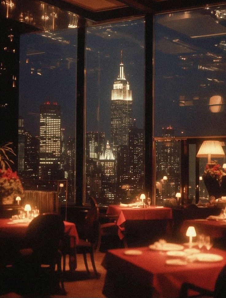 Restaurant with large windows showing New York City skyline at night with Empire State Building, candlelit tables in foreground