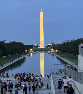 Washington Monument lit up at dusk with its reflection in the Reflecting Pool and crowds of visitors