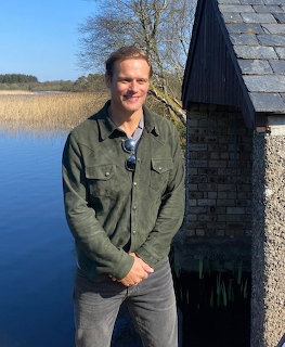Man standing by a lakeside next to a stone building on a sunny day