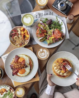 Flatlay of brunch dishes including eggs, salmon salad, granola bowl, and coffee on a cafe table with a camera