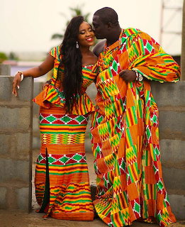 Couple in colorful kente cloth wedding attire, groom kissing bride's forehead outdoors
