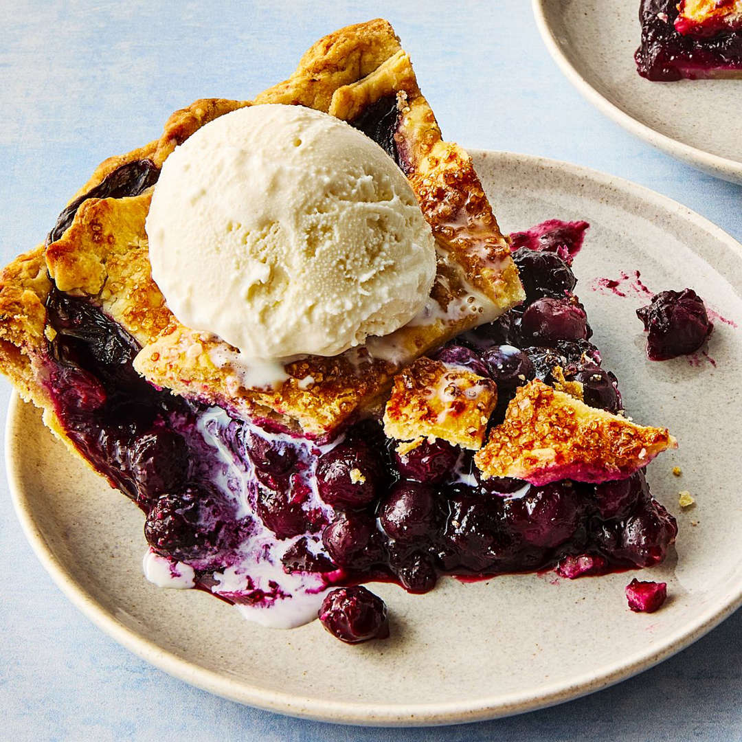 Slice of blueberry pie with lattice crust and vanilla ice cream scoop on white plate