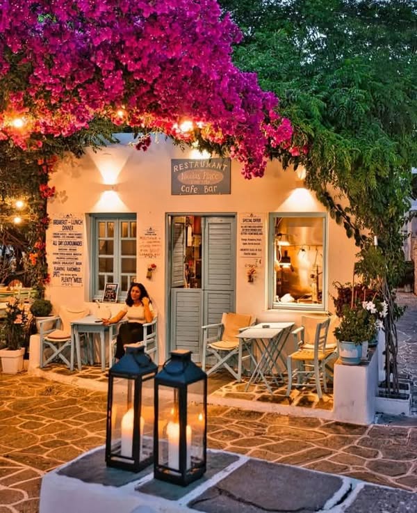 Whitewashed Greek island café at dusk with pink bougainvillea, candlelit lanterns, and outdoor seating on a stone terrace.