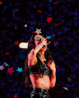 Olivia Rodrigo performing on stage with a microphone and colorful star lights in the background.