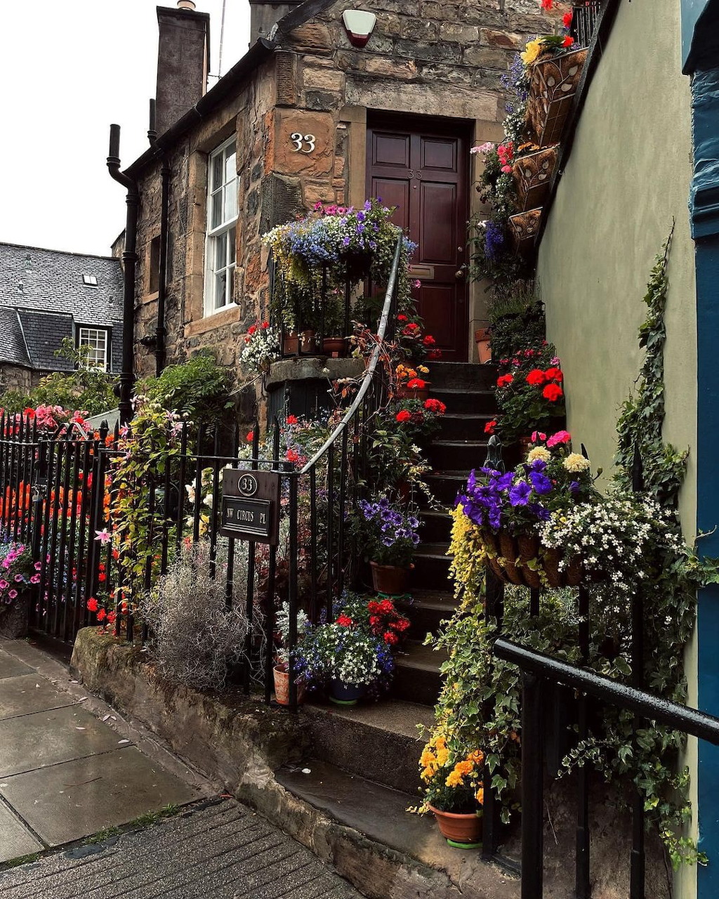 Stone cottage entrance with steps covered in colorful poted flowers and hanging baskets along a narrow street