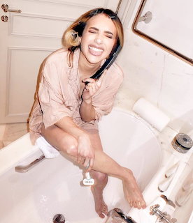 Young woman laughing and holding a hairbrush while sitting in a white bathtub wearing a pink shirt