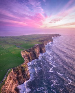 Aerial view of dramatic coastal cliffs with green fields meeting ocean under pink and purple sunset sky