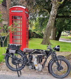Black vintage 1928 Sunbeam motorcycle beside red telephone box on street with green lawn and trees