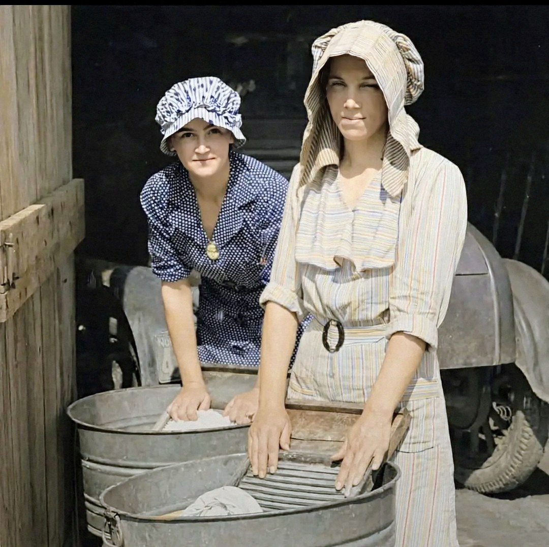 Two women washing clothes by hand in metal tubs with washboards in a vintage colorized photo