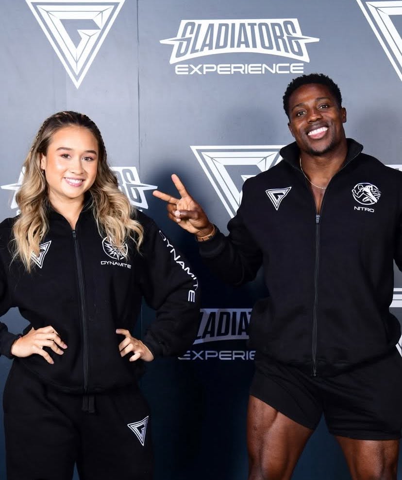 Two smiling athletes in black tracksuits pose in front of a Gladiators Experience backdrop
