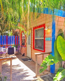 Pink cottage exterior with red-trimed window, blue fence, tropical plants, and woodenorch