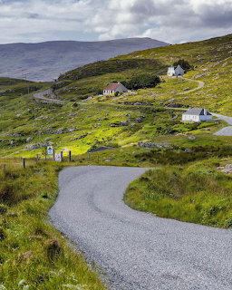 Winding road through green hillside with white cottages and distant mountains under cloudy skies