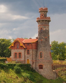 Stone dam keeper's house with a tall tower and red roof, surrounded by greenery under cloudy skies