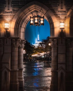 Castle lit up at twilight, framed by gothic archway with lanterns and wet stone pathway