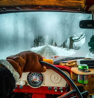 Interior of vintage truck with gloved hand on wheel, looking out at snowy forest road through foggy windshield