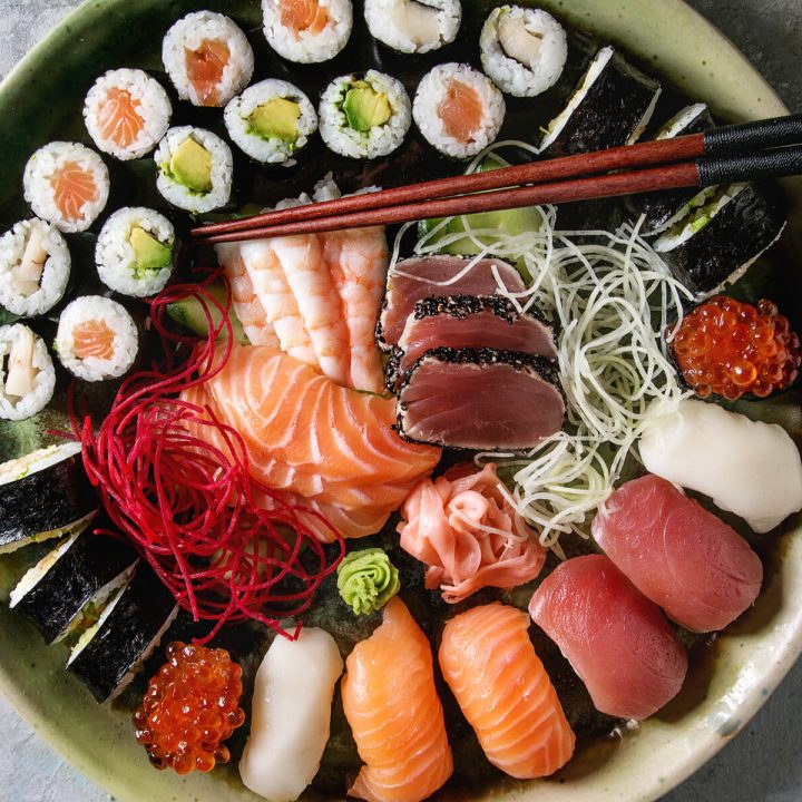 Overhead view of assorted sushi platter with nigiri, sashimi, and maki rolls on dark plate with chopsticks