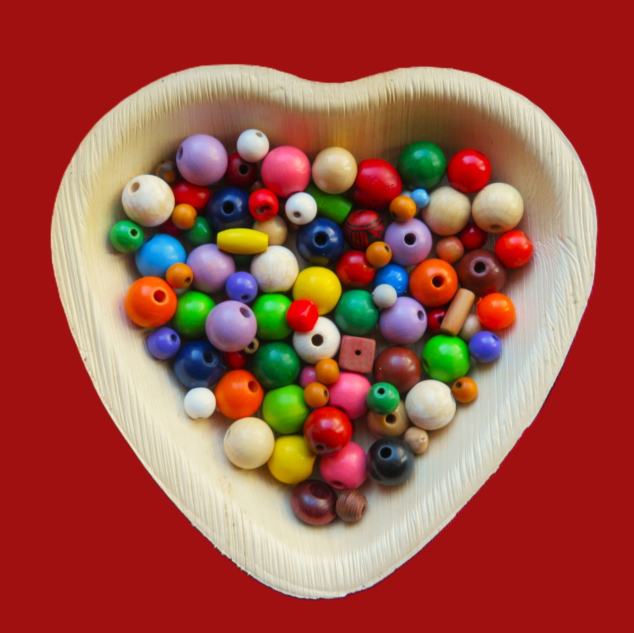Colorful wooden beads in a heart-shaped wooden bowl on a red background