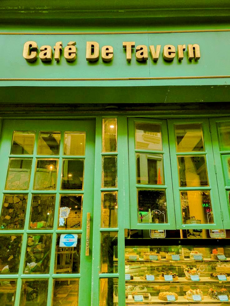 Green facade of Café De Tavern with large windows displaying pastries and baked goods