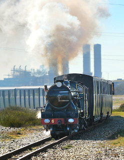 Black steam train on tracks with large white steam cloud and power station cooling towers behind