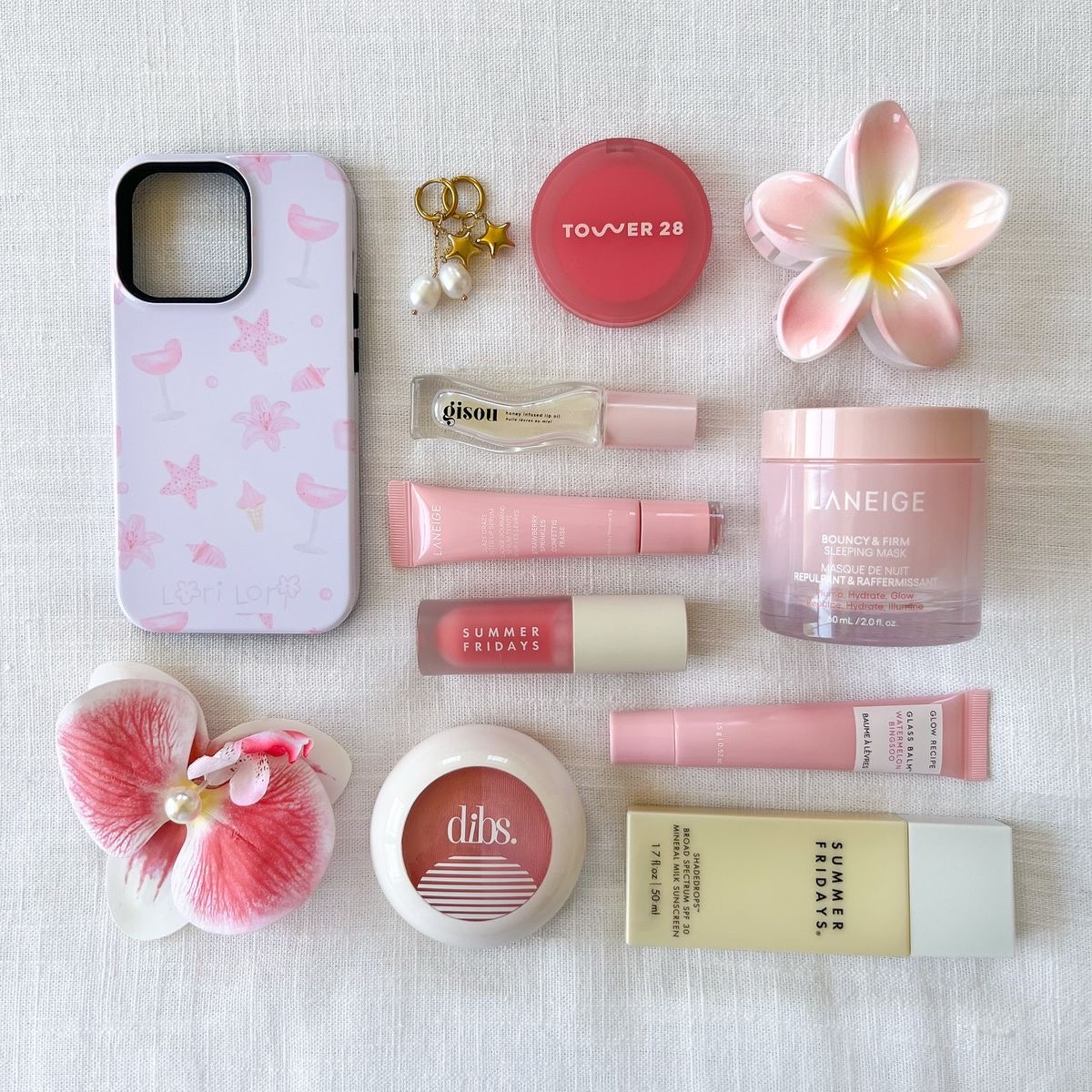 Flat lay of pink makeup products, skincare items, phone case with flower clips, and plumeria blossom on white marble