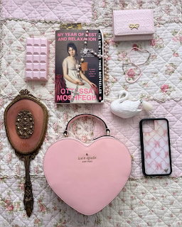 Pink flat lay on a floral quilt with a heart-shaped box, hand mirror, phone, wallet, ceramic bird, and magazine