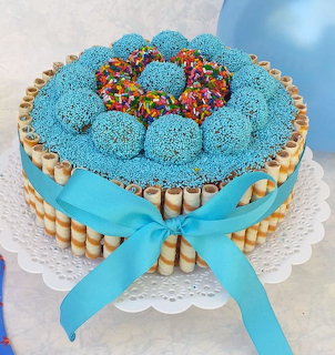 Round cake covered in blue sprinkles with colorful candies on top, wafer cookie border, and blue ribbon bow