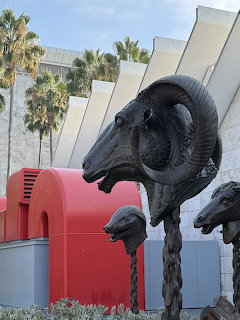 Bronze ram head sculptures with large curved horns in front of a modern building with palm trees