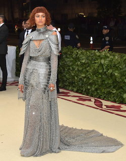 Zendaya standing on a red carpet in a silver chainmail-style gown with a long train