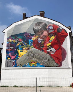 Street mural of child with colorful toys painted on white building exterior under blue sky