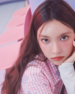 Young woman with long brown hair in pink gingham shirt posing with hand on chin against pastel background