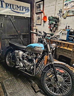 Blue and chrome Triumph motorcycle parked in cluttered vintage workshop with tools and signs on wooden walls